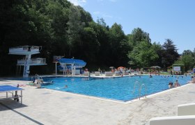 Ein Freibad mit Sprungturm, Rutsche und vielen Badegästen an einem sonnigen Tag.