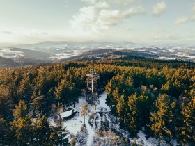 L&eacute;gi felv&eacute;tel egy h&oacute;val bor&iacute;tott tűlevelű erdőről, a h&aacute;tt&eacute;rben egy kil&aacute;t&oacute;toronnyal &eacute;s dombokkal.