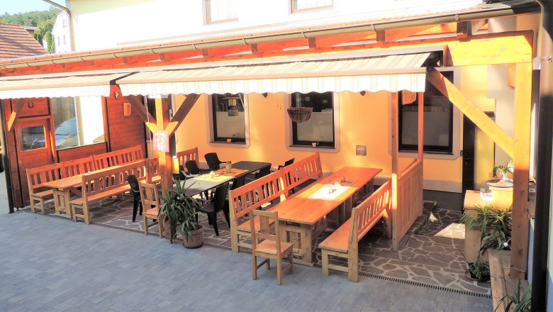 Ein gemütlicher Gastgarten mit Holzbänken und Tischen unter einer Markise.