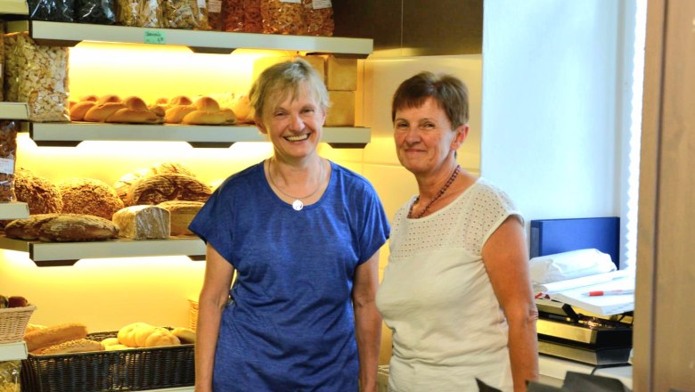 Zwei Frauen in einer Bäckerei vor Regalen mit Brot und Gebäck.