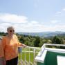 Elderly woman with sunglasses stands on a balcony overlooking a hilly landscape.