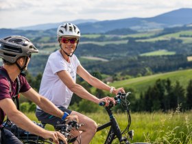 Zwei Radfahrer mit Helmen fahren durch eine gr&uuml;ne Landschaft mit H&uuml;geln im Hintergrund.