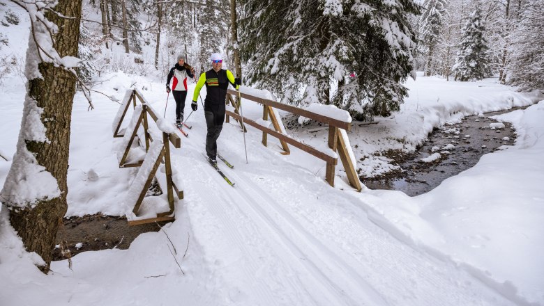 Zwei Personen &uuml;berqueren beim Langlaufen einen Bach in einer verschneiten Waldlandschaft
