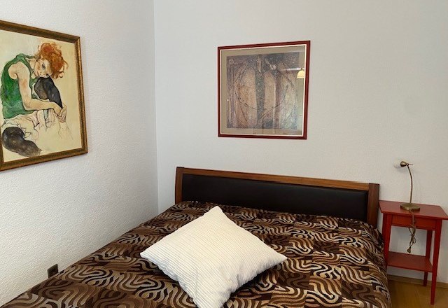 A bedroom with a bed, two framed pictures on the wall and a red bedside table.