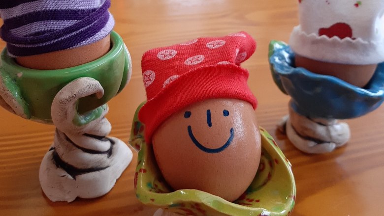 Drei Eier in bunten Eierbechern, eines mit einem aufgemalten Gesicht und roter Mütze.