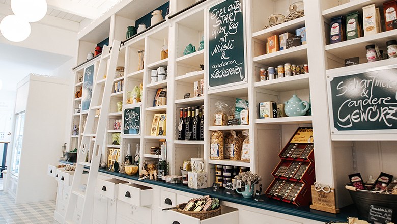 Hofladen mit weißen Holzregalen und diversen Produkten wie Gewürzen, Schokoladen und Tees.