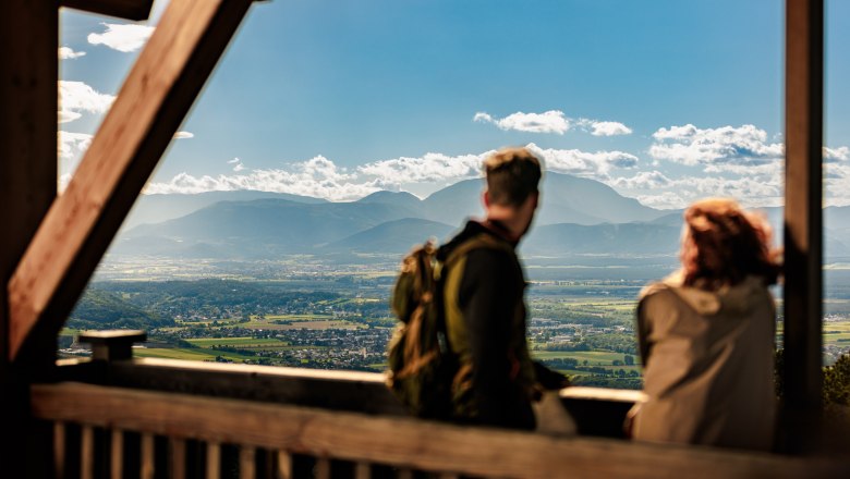Zwei Personen stehen auf der Plattform des hölzernen Aussichtsturmes Lanzenkirchen/Wiesen, im Hintergrund ist eine weite Ebene und am Horizont der Schneeberg gut erkennbar.