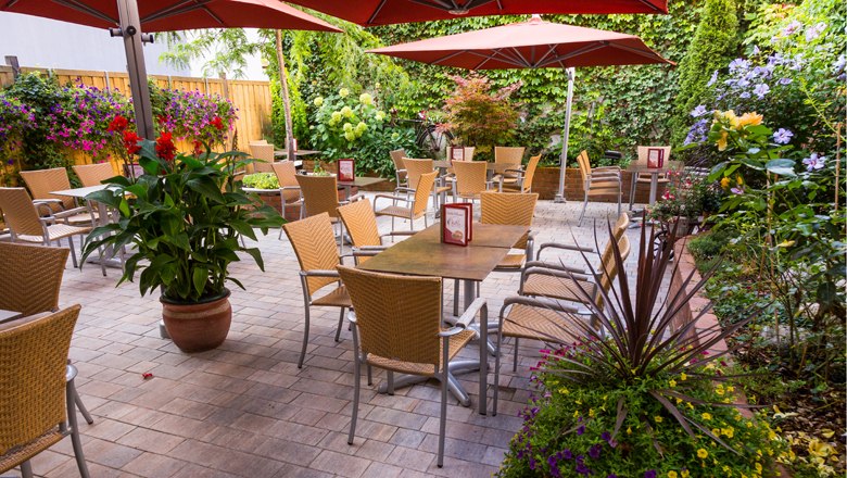 Ein gemütlicher Gastgarten mit Tischen, Stühlen und Sonnenschirmen, umgeben von üppiger Bepflanzung.
