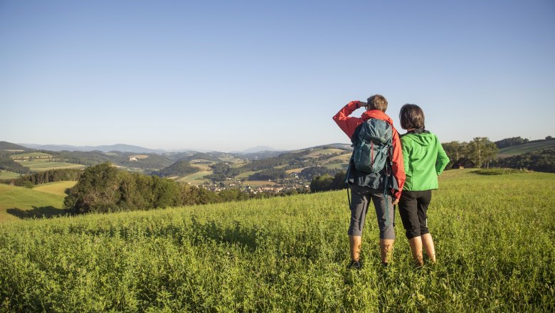 Zwei Personen stehen auf einer Wiese und blicken in die Ferne über eine hügelige Landschaft.