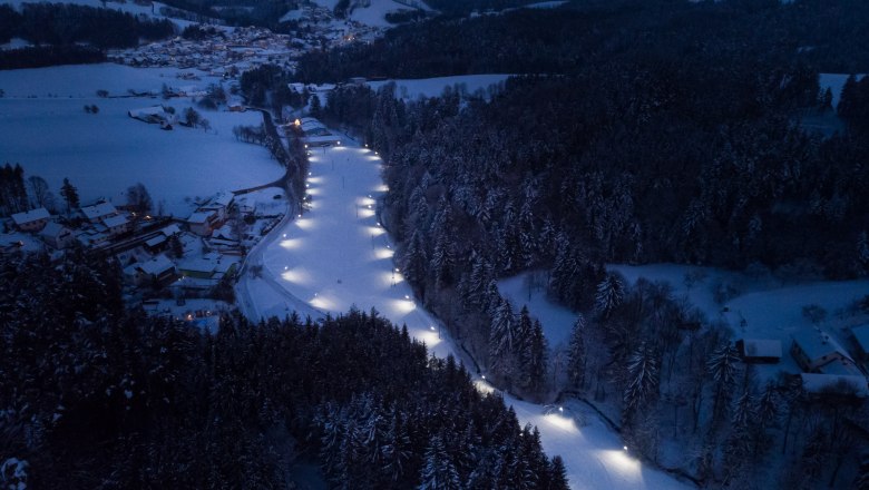 Luftaufnahme einer beleuchteten Langlaufloipe in einer verschneiten Landschaft bei Nacht.