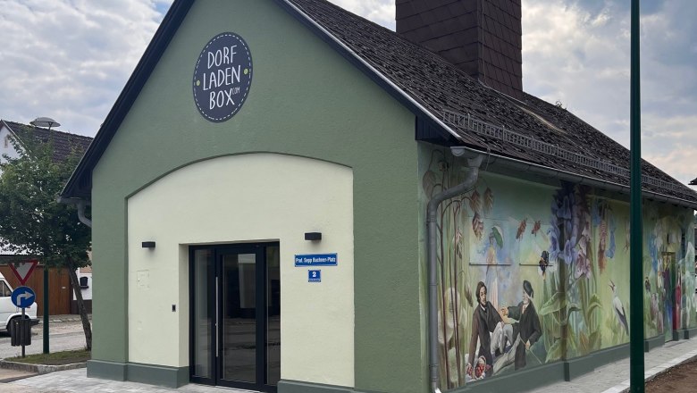 A small, green building with the inscription 'Dorfladen Box' and a painted, rural mural.