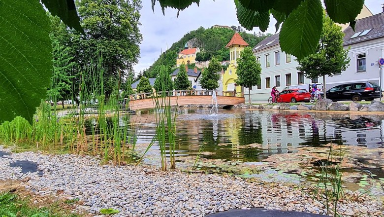 Blick auf einen Teich mit Brücke und Bergkirche im Hintergrund, umgeben von Bäumen und Häusern.
