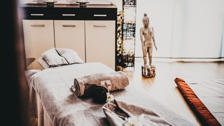 Ein Spa-Raum mit Massageliege, Handt&uuml;chern, Buddha-Statue und Kerzen.