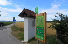 Radstartplatz-Tafel in Katzelsdorf mit Wegweiser und Landschaft im Hintergrund.