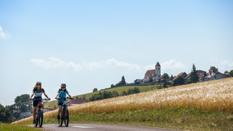 Zwei Personen fahren mit E-Bikes auf einem ländlichen Weg, im Hintergrund ein Dorf mit Kirche.