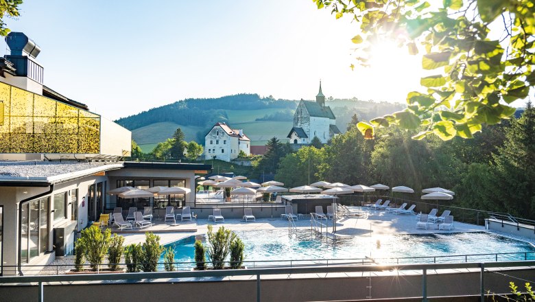 Vivea 4* Hotel Bad Schönau Zum Landsknecht, © Hannes Dabernig Außengelände eines Hotels mit Pool, Sonnenschirmen und Liegen, umgeben von Natur und Kirche im Hintergrund.