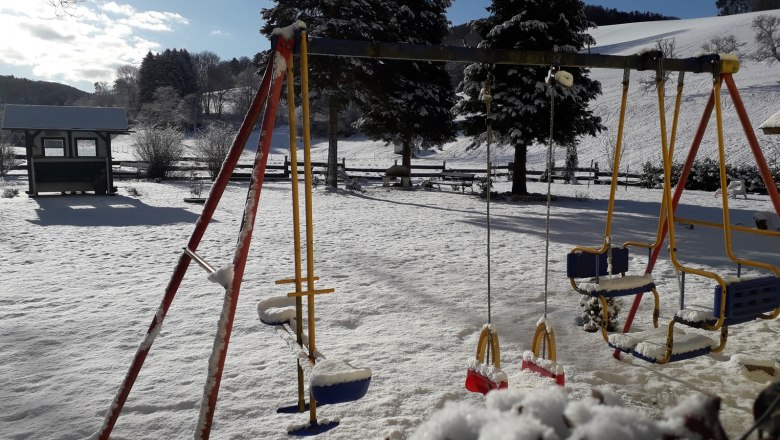 Schneebedeckter Spielplatz mit Schaukeln und Bäumen im Hintergrund.