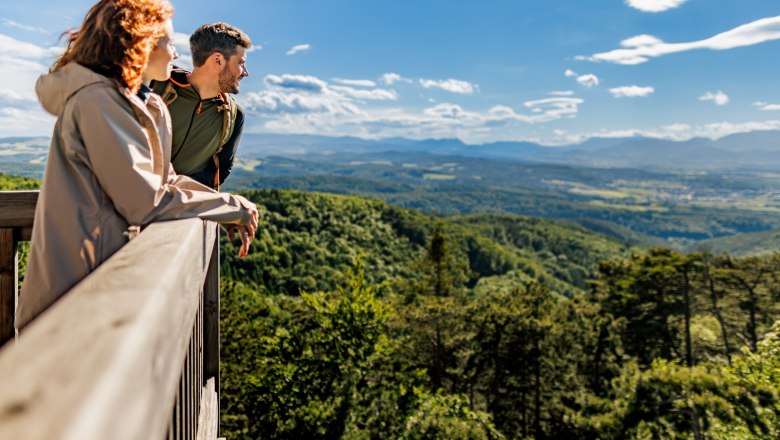 Zwei Personen stehen auf der Plattform des Aussichtsturmes Lanzenkirchen/Wiesen und blicken über eine bewaldete Landschaft mit Bergen im Hintergrund.