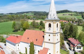 Luftaufnahme der Wehrkirche Krumbach in einer l&auml;ndlichen Umgebung mit gr&uuml;nen Feldern und blauem Himmel.