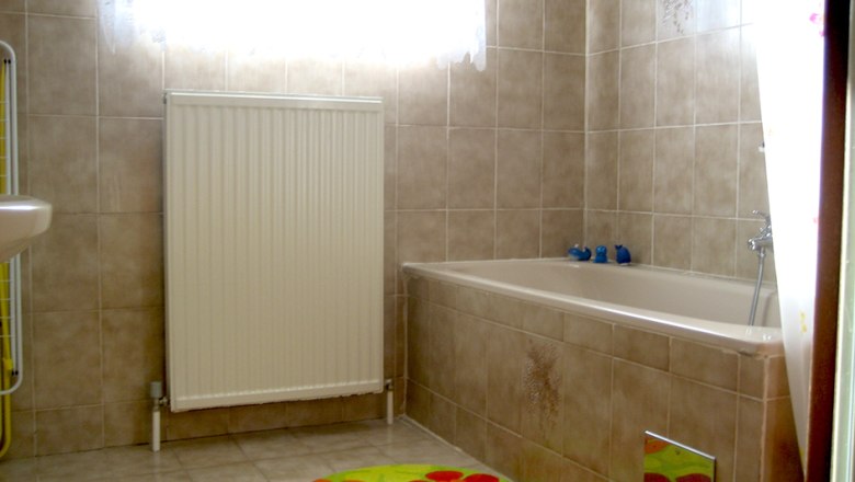 A bathroom with beige tiled wall, bathtub, radiator and colorful carpets.