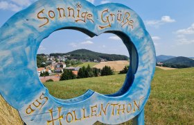 Blauer Holzrahmen in Form einer Wolke mit Schriftzug 'Sonnige Grüße aus Hollenthon' vor Landschaft.