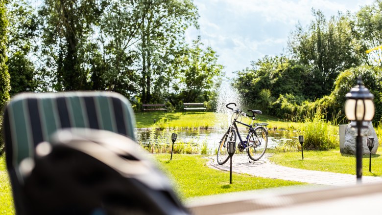 Ein Fahrrad steht neben einem Teich mit Springbrunnen in einem sonnigen Garten.