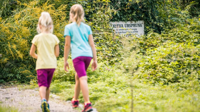 Zwei Kinder gehen auf einem Weg in einem grünen Waldgebiet. Ein mit Efeu bewachsenes Schild mit der Aufschrift 'Leitha Ursprung' ist im Hintergrund sichtbar.