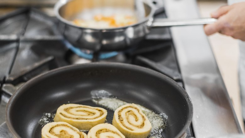 Egy serpenyő h&aacute;rom porcini gomb&aacute;s rol&oacute;val a tűzhelyen, mellette egy k&eacute;z, amely egy m&aacute;sik serpenyőt tart.