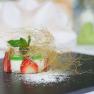 An artfully arranged goat's cheese tartlet with strawberries and mint, wrapped in caramel threads, on a black plate.