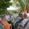 Elderly people sit around a table on a terrace, surrounded by trees and with a view of the countryside.