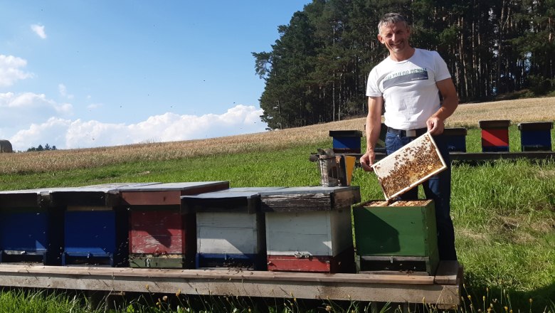 Ein l&auml;chelnder Mann mit einer Honigwabe in der Hand steht neben einer Reihe von Bienenst&ouml;cken.