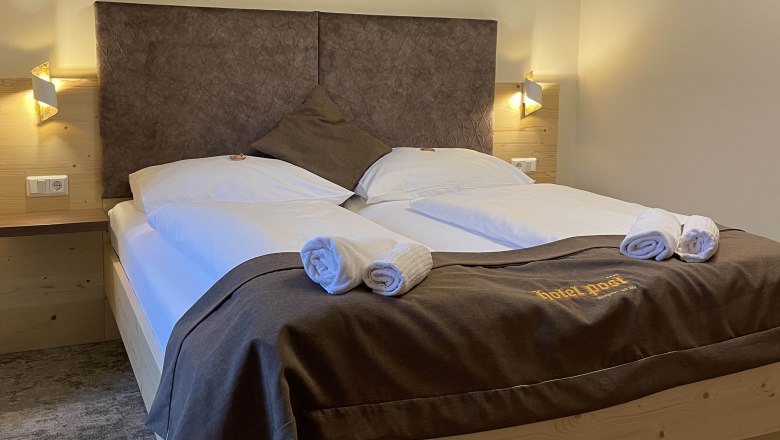 Ein gemütliches Doppelzimmer mit großem Bett, dekoriert mit brauner Bettwäsche und Handtüchern. An der Wand sind geometrische Muster.