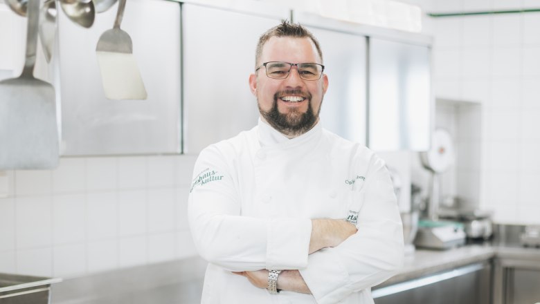 A smiling chef in a white uniform stands in a professional kitchen.