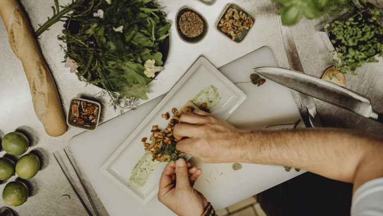 Chef decorating a dish with herbs on a plate in a kitchen.