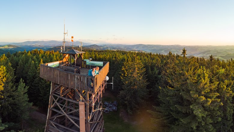 Aussichtsturm aus Holz im Wald mit zwei Personen, die den Sonnenuntergang betrachten, im Hintergrund hügeliges Panorama,