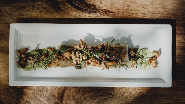 A rectangular plate with saut&eacute;ed chanterelles, pecan nuts, wild herbs and root bread, artfully arranged on a wooden table.