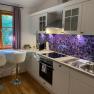 Modern kitchen with white cupboards, lavender splashback and two bar stools in front of a window with purple curtains.