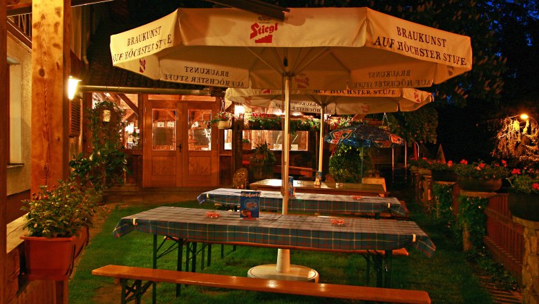 Ein gemütlicher Biergarten bei Nacht mit Holztischen, blau karierten Tischdecken und großen Sonnenschirmen der Marke Stiegl.