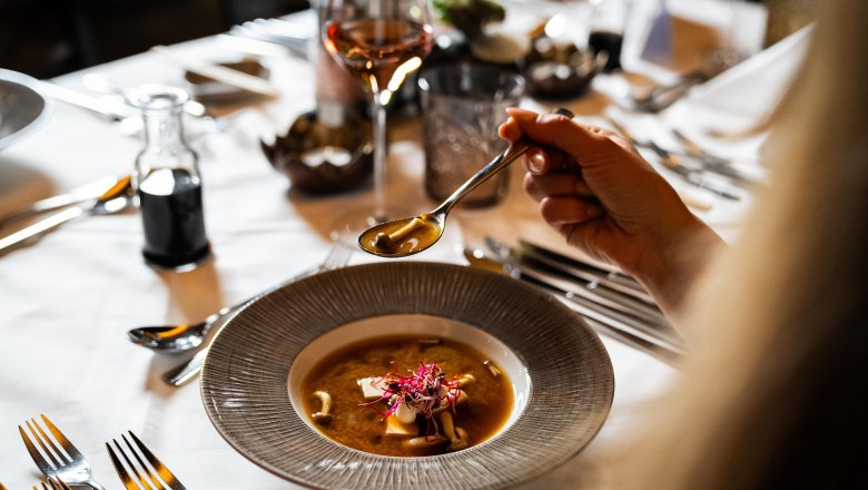 Eine Person hält einen Löffel über einem Teller mit Suppe auf einem gedeckten Tisch.