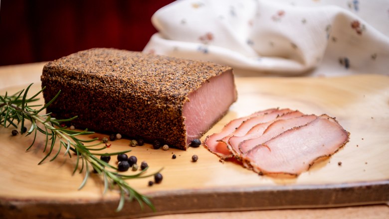 Ein Stück gepökeltes Fleisch mit Pfefferkruste und geschnittenen Scheiben auf einem Holzbrett, dekoriert mit Rosmarin und Wacholderbeeren.