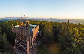 Aussichtsturm aus Holz im Wald mit zwei Personen, die den Sonnenuntergang betrachten, im Hintergrund hügeliges Panorama,