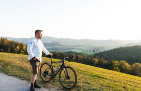 Ein Mann in wei&szlig;em Hemd steht mit einem Fahrrad auf einem H&uuml;gel mit Blick auf eine gr&uuml;ne Landschaft.