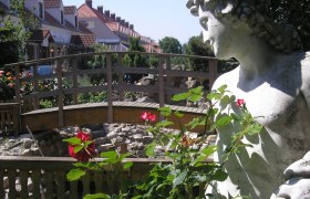 Steinstatue neben einer Holzbr&uuml;cke und roten Rosen im Vordergrund.