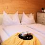 Cozy bedroom with double bed, white pillows and blankets, yellow blanket, tray with cups, bedside lamp and chest of drawers with towels.