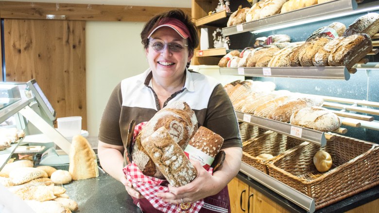 Eine Frau in einer Bäckerei hält mehrere Brote in den Händen und lächelt in die Kamera.