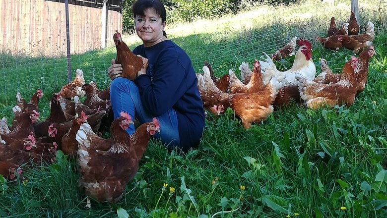 Eine lächelnde Frau hält ein Huhn auf einer grünen Wiese, umgeben von vielen weiteren Hühnern.