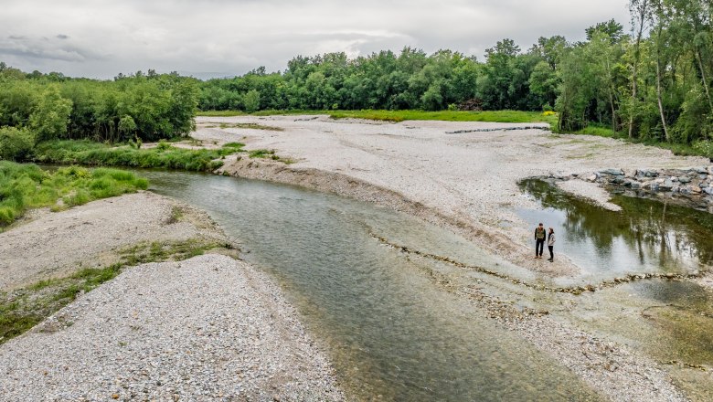 Ein weites Schotterbett eines Flusses durch das klares Wasser fließt. Zwei Personen stehen am Fluss, im Hintergrund grüner Wald