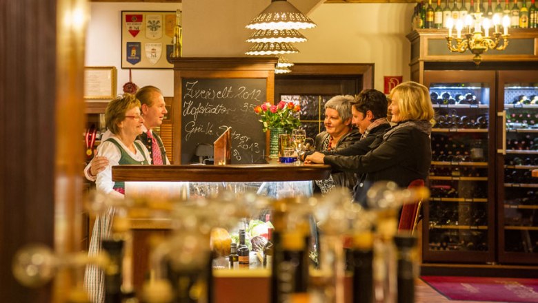 Menschen in einer gemütlichen Gaststube mit Weinflaschen im Hintergrund.