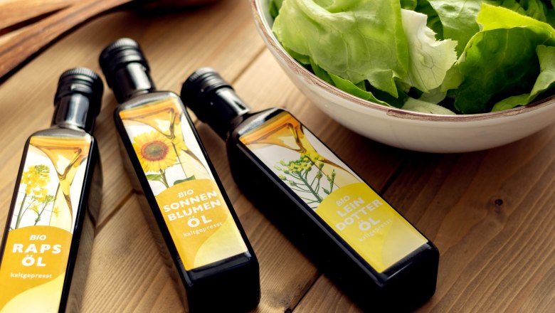 Drei verschiedene &Ouml;le liegen auf einem Holztisch neben einer Sch&uuml;ssel mit gr&uuml;nem Salat.